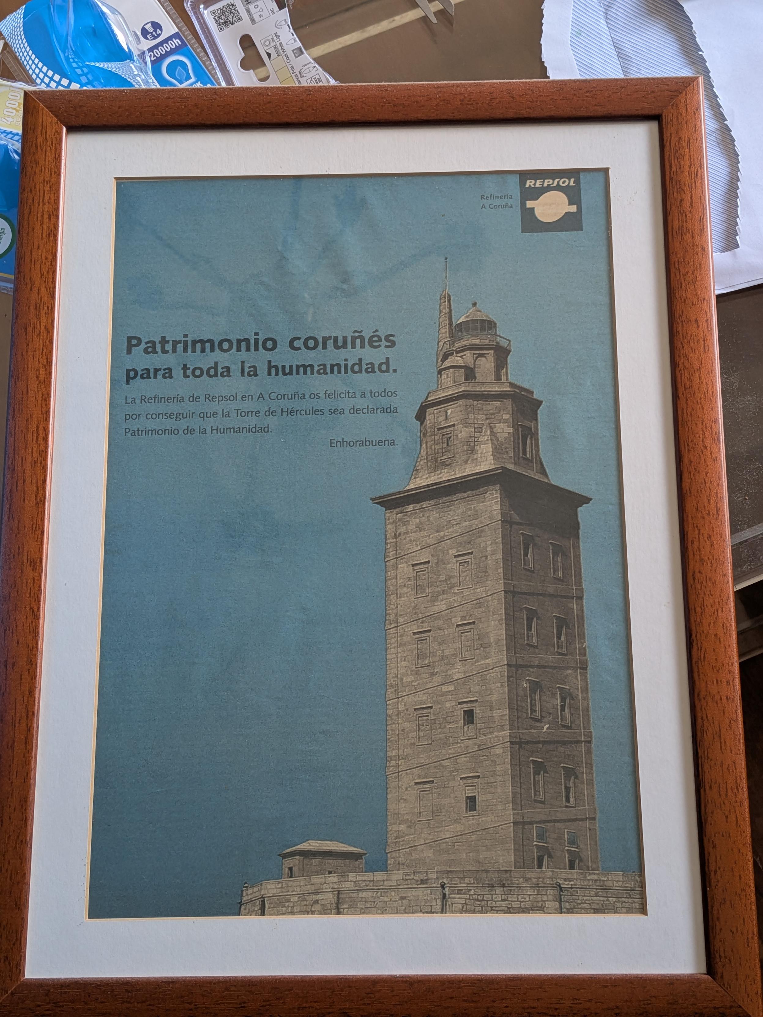 Un cuadro con una fotografía de la torre de Hércules que dice Patrimonio coruñés para toda la humanidad en letras oscuras en el cielo. Arriba se ve el logo de Repsol.